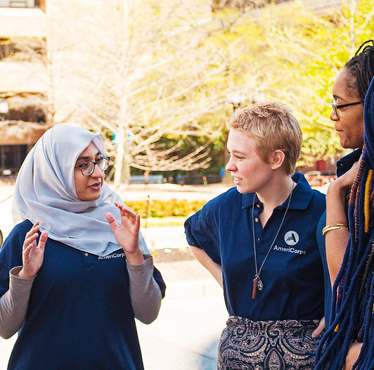 Group of AmeriCorps VISTA members talking, including a young woman in a head scarf and glasses, a young white woman with her hands on her hips, and a young black woman wearing glasses